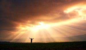 A person with their arms raised in praise silhouetted against the golden rays from a partly clouded sky.